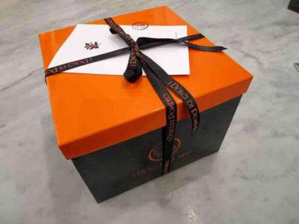 Una scatola nera e arancio con il marchio "I dolci di Pompeo" in rilievo legata con un nastro di raso nero.